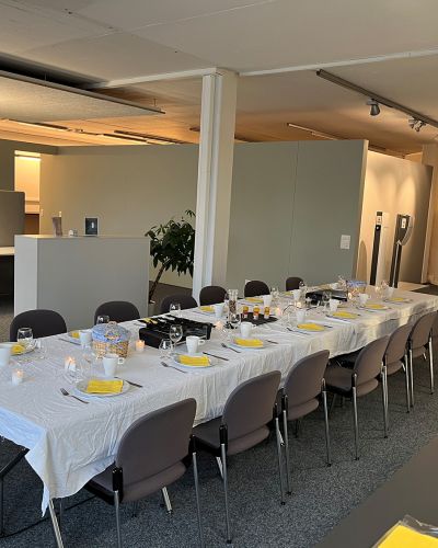 Ein schön gedeckter Tisch mit 14 Stühlen in unseren neuen Räumlichkeiten. Alles ist für einen feierlichen Raclette-Abend vorbereitet, um die Einweihung unserer neuen Räumlichkeiten zu feiern.
